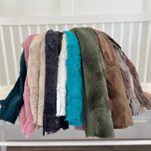 May include: A collection of plush, faux fur fabric in various colors, including teal, pink, beige, navy, white, turquoise, olive green, brown, and a patterned brown and gray. The fabrics are draped over a white bench, showcasing their soft texture.
