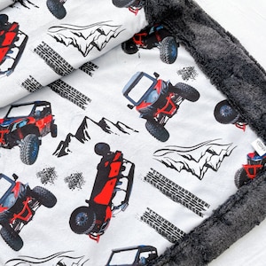 May include: A white fleece blanket with a red and black off-road vehicle pattern. The blanket has a gray fleece backing.