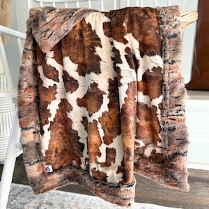 May include: A plush throw blanket featuring a brown and white cowhide pattern with a textured, shaggy border. The fabric has a soft, faux fur appearance with irregular patches of dark brown, light brown, and white throughout the design.
