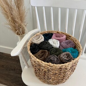 May include: A woven basket filled with rolled, colorful faux fur fabric. The basket sits on a white chair. The fur rolls are in shades of teal, pink, purple, brown, black, and white. A vase with dried grass is in the background.