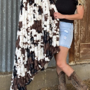 May include: A plush blanket with a cowhide pattern in shades of brown, black, and white. The faux fur blanket is held up, showcasing its soft texture and design. The blanket is a cozy home decor item.