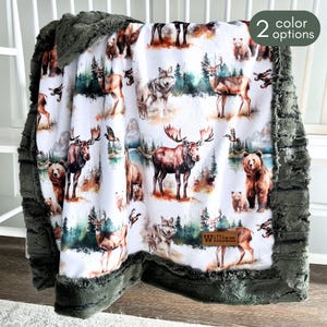 May include: A white baby blanket with a woodland animal print featuring moose, deer, bears, and wolves. The blanket has a green faux fur trim and the name "William" is embroidered on the bottom right corner.