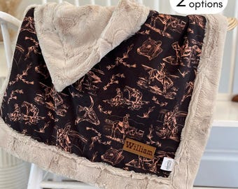 May include: A black and white baby blanket with a brown faux fur border. The blanket features a repeating pattern of cowboys, horses, and cattle. The blanket has a personalized label with the name "William" on it.