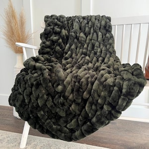 May include: A dark green, textured faux fur throw blanket draped over a white chair. The blanket has a plush, dimensional appearance, with a woven pattern. The background includes a white wall and a decorative vase with dried plants.