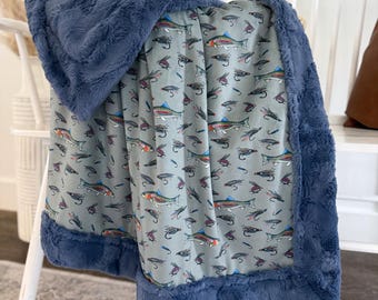 May include: A soft, blue blanket with a fish and fly pattern. The blanket has a plush, navy blue border. The blanket is draped over a white chair. The blanket's pattern features colorful fish and fishing flies on a light gray background.