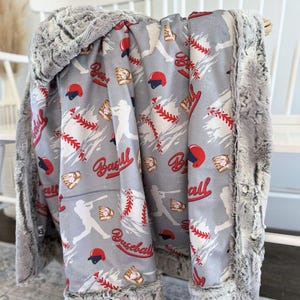 May include: A gray baseball-themed blanket with a soft, plush border. The blanket features baseballs, helmets, gloves, and baseball players in red, white, and gray. The blanket is draped over a white chair.