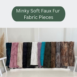 May include: A display of Minky Soft Faux Fur Fabric Pieces in various colors, including teal, pink, beige, black, white, purple, and brown. The fabric has a soft, textured appearance, and the pieces are arranged on a white shelf.