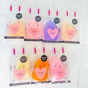 May include: Six colorful heart-shaped makeup sponges in clear packaging with bunny ears. Each sponge has a cute cartoon face and the words "Make Up Sponge" and "beauty" printed on it. The packaging says "High Quality Material".