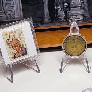 May include: A clear acrylic display stand with a triangular base and two angled supports. The stand is positioned in the foreground, while three other stands hold a Walt Disney postage stamp, a gold-toned coin, and a gold-toned castle plate.