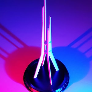 May include: A white, geometric model of a tall, slender tower with a black base. The model is lit with pink and blue lights, casting shadows on the surface.