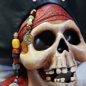 May include: A human skull replica wearing a black leather pirate hat with a white bone cross emblem. A red patterned bandana is wrapped around the skull, adorned with a strand of multi-colored beads and a circular bronze lion pendant.
