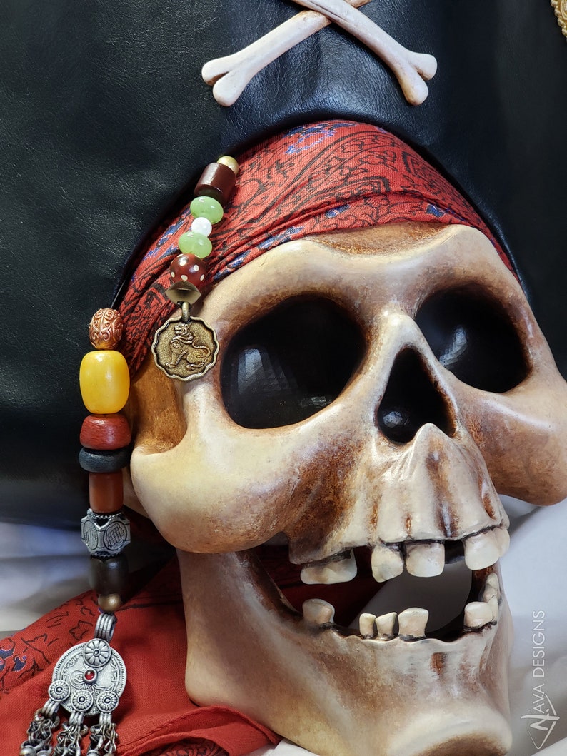 May include: A human skull replica wearing a black leather pirate hat with a white bone cross emblem. A red patterned bandana is wrapped around the skull, adorned with a strand of multi-colored beads and a circular bronze lion pendant.