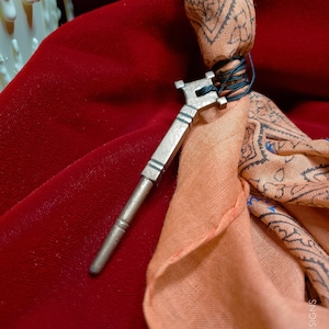 Puede incluir: Una llave plateada con una parte superior decorativa, atada con un cordón negro a una tela de color melocotón con un patrón de paisley. La tela está sobre una superficie de terciopelo rojo. *Muestra únicamente. No viene prepintado ni premontado.*
