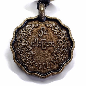 May include: A circular bronze-colored pendant with a scalloped edge and a textured surface. The center features embossed script characters surrounded by an intricate floral border. A black cord passes through a loop at the top of the pendant.