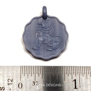 May include: A circular gray pendant with a scalloped edge featuring an embossed mythical lion figure. The pendant is positioned above a metal ruler showing a diameter of approximately 1 inch. The surface has a matte finish with visible texture.