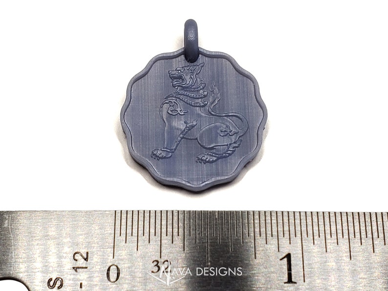 May include: A circular gray pendant with a scalloped edge featuring an embossed mythical lion figure. The pendant is positioned above a metal ruler showing a diameter of approximately 1 inch. The surface has a matte finish with visible texture.