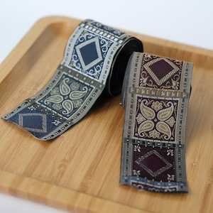 Könnte beinhalten: Zwei Rollen gewebtes Jacquardband mit komplizierten geometrischen Rautenmustern und stilisierten Paisley-Motiven. Ein Band zeigt Schattierungen in Marineblau und Grau, während das andere in tiefen Burgunder- und Goldtönen mit metallischen Akzenten gehalten ist.