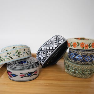 Op de afbeelding: Een collectie van zes decoratieve stoffen linten opgerold tot spoelen, met diverse patronen waaronder bloemmotieven, geometrische vormen en ingewikkeld borduurwerk in kleuren zoals zwart, wit, blauw, oranje, groen en metallic gouden draden.