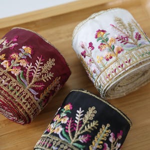 Könnte beinhalten: Drei Rollen dekoratives Stoffband in Burgunderrot, Weiß und Schwarz. Jede Rolle weist eine aufwendige Blumenstickerei mit goldenen Metallic-Fadenakzenten und kleinen Pailletten an den Rändern auf, die detaillierte botanische Muster zeigen.