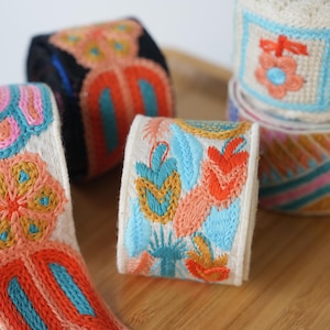 Peut inclure: Plusieurs rouleaux de ruban décoratif en tissu avec des broderies complexes dans des tons vifs d'orange, bleu, rose et jaune. Les motifs incluent des fleurs, des formes géométriques et des feuilles stylisées sur des bases en tissu crème et noir.