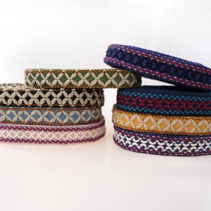 Könnte beinhalten: Eine Sammlung farbenfroher, gemusterter Bänder, die übereinander gestapelt sind. Die Bänder weisen verschiedene Designs in Blau-, Grün-, Lila- und Goldtönen auf. Die Bänder scheinen etwa 2,5 cm breit zu sein.