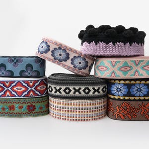 Puede incluir: Una colección de nueve rollos de cinta y pasamanería tejida decorativa con varios patrones, incluyendo motivos florales, formas geométricas y rayas en colores como azul, rojo, verde, rosa, negro y marrón, con un rollo rematado con pompones negros.