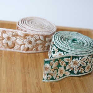 Könnte beinhalten: Zwei Rollen dekoratives Stoffband mit aufwendiger Blumenstickerei. Eine Rolle ist in einem sanften Beigeton gehalten, die andere in einem tiefen Waldgrün. Beide Bänder zeigen ein wiederkehrendes Muster aus weiß und gold gestickten Blüten.