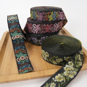 Könnte beinhalten: Mehrere Rollen schwarzes Stoffband mit komplizierten, gewebten Blumenmustern in leuchtenden Farben wie Rot, Rosa, Violett, Orange, Grün und Gelb. Die Bänder sind als aufgerollte Spulen dargestellt, wobei ein Ende jedes Bandes nach außen verläuft.