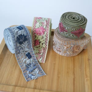 Op de afbeelding: Drie rollen decoratief geborduurd lint met ingewikkelde bloemmotieven. Eén rol is lichtblauw met donkerblauwe bloemen, één is lichtgroen met roze bloemen en één is neutraal beige met bruin bloemenborduurwerk.