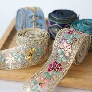 Könnte beinhalten: Fünf Rollen dekoratives Stoffband mit aufwendiger Blumenstickerei und Pailletten. Die Bänder zeigen verschiedene Farben wie Gold, Rosa, Blaugrün und Blau, mit metallisch goldenen, gewellten Kanten auf transparentem Netzstoff.