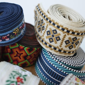Op de afbeelding: Een verzameling rollen stoffen lint met diverse geweven patronen. Eén rol is marineblauw met bloemendetails, een andere is crème met gouden en zwarte geometrische vormen, en andere tonen levendige neon oranje, groene en blauwe ontwerpen.