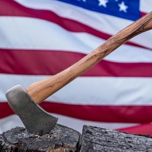 May include: A wood-handled axe with a silver blade is embedded in a tree stump. The axe handle has a natural wood grain pattern. The background features the American flag, with its red and white stripes and blue field of stars.