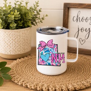 May include: A white ceramic mug with a handle and a clear lid featuring a graphic of the state of New Mexico. The design includes a pink and white checkered bow, a blue heart, and the text New Mexico and NM in bright pink and blue colors.