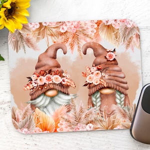 May include: A mouse pad featuring two gnomes with brown hats and floral crowns. The gnomes are surrounded by beige and orange palm leaves and flowers.