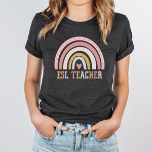 May include: A dark charcoal gray short-sleeve t-shirt featuring a graphic of a multi-colored rainbow in shades of pink, yellow, and blue with a small heart at the center. The text ESL TEACHER is printed in block letters below the rainbow graphic.