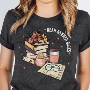 May include: A dark charcoal gray t-shirt featuring a graphic print of a stack of books topped with flowers, a pink mug with whipped cream, a coffee cup, and an open book with round glasses. The text READ BANNED BOOKS is printed in white.