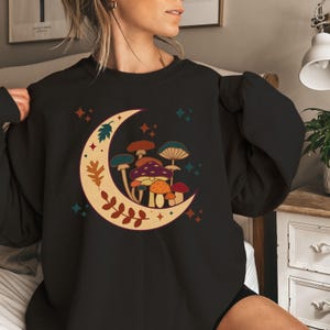 May include: A black crewneck sweatshirt featuring a graphic of a crescent moon filled with various mushrooms in shades of orange, purple, and blue. The moon design is decorated with leaf motifs and small four-pointed stars in teal and orange colors.