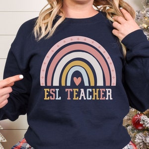 May include: A navy blue long-sleeve sweatshirt featuring a graphic of a multi-colored rainbow with a small heart at the center and the text ESL TEACHER printed in block letters below. The rainbow design includes small star accents along the top arch.