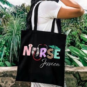 May include: A black tote bag featuring the word NURSE in large, multi-patterned letters, including pink hearts, plaid, purple medical icons, leopard print, and teal stripes. A red stethoscope and a green caduceus symbol are integrated. The name Jessica is printed below in white script.