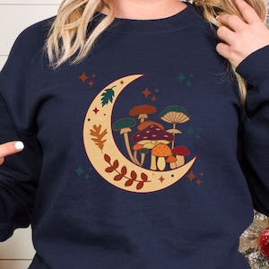 May include: A navy blue crewneck sweatshirt featuring a graphic print of a large cream-colored crescent moon decorated with brown leaves and a cluster of various mushrooms in shades of orange, red, brown, and green, accented by small star shapes.