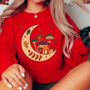 May include: A bright red long-sleeve sweatshirt featuring a large graphic of a cream-colored crescent moon filled with various brown, orange, and green mushrooms, accented by small star shapes and leaf patterns printed on the front center.