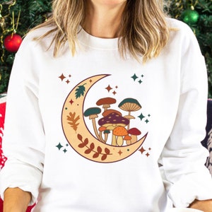 May include: A white long-sleeve sweatshirt featuring a graphic print of a crescent moon decorated with leaves and a cluster of various mushrooms in shades of brown, orange, and teal, accented by small four-pointed star shapes in dark green and rust.