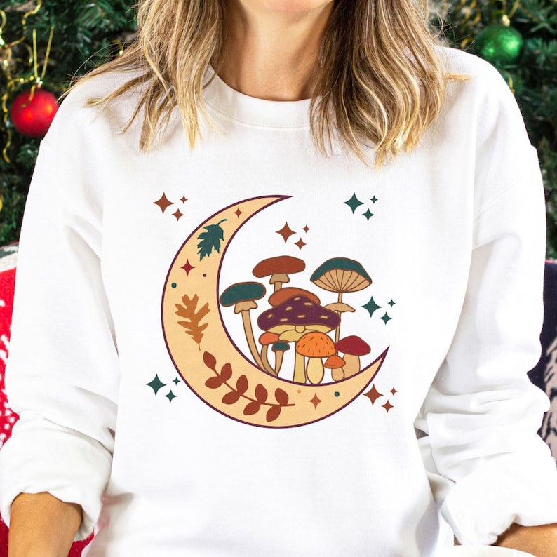 May include: A white long-sleeve sweatshirt featuring a graphic print of a crescent moon decorated with leaves and a cluster of various mushrooms in shades of brown, orange, and teal, accented by small four-pointed star shapes in dark green and rust.