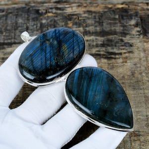 May include: Two labradorite pendants with silver-colored settings. One is oval, the other teardrop-shaped. The stones have a dark blue and black iridescence with streaks of yellow and green. The pendants are displayed on a white surface.