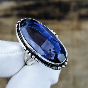 May include: A silver ring featuring a large, oval-cut, deep blue gemstone. The metal setting has a decorative beaded border along the edges of the oval frame, and the band is split into two sections where it connects to the main stone setting.