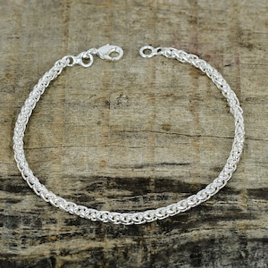 Peut inclure: Un bracelet en argent avec un motif de chaîne tressée. Le bracelet est doté d'une fermeture à fermoir mousqueton. Le bijou est présenté sur une surface en bois. C'est un bijou délicat.