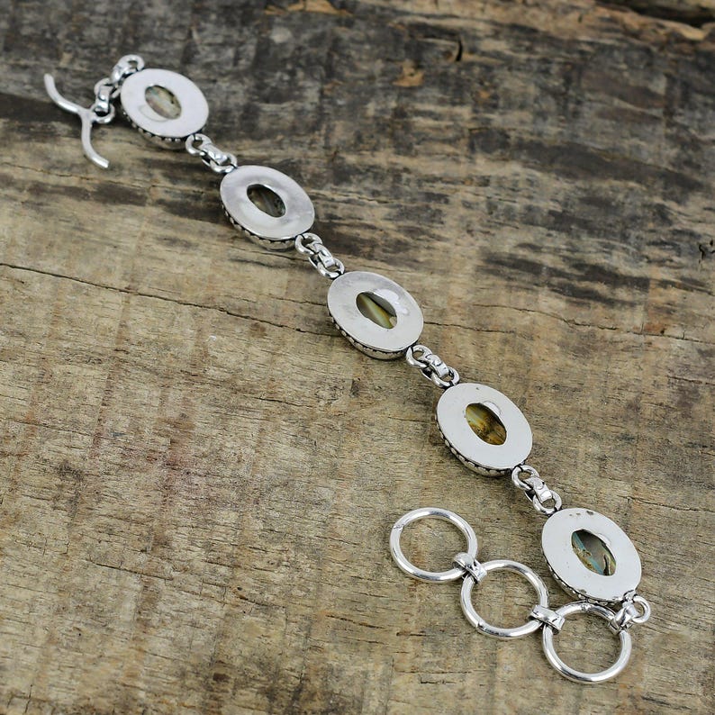 K&ouml;nnte beinhalten: Ein silbernes Armband mit ovalen Steinen in einem Ketten-Design. Das Armband hat einen Hakenverschluss und eine Reihe von Verbindungsringen. Die Steine haben einen hellgr&uuml;nen Farbton, und das Metall ist poliert.