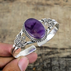 May include: A silver cuff bracelet with an oval, deep purple amethyst gemstone. The bracelet features butterfly and floral designs. The gemstone is set in a detailed bezel setting. The bracelet is a beautiful piece of jewellery.