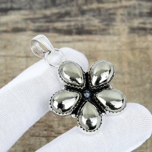 May include: Silver-toned pendant in the shape of a flower. The pendant features five teardrop-shaped petals with a metallic, reflective surface. A small, light blue stone is at the center. The pendant is held by a silver loop.
