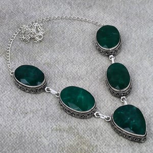 Pode incluir: Um colar de prata com cinco pedras preciosas ovais, facetadas, verde esmeralda. As pedras são engastadas em biséis prateados ornamentados. O colar tem uma corrente delicada e é exibido em uma superfície cinza.